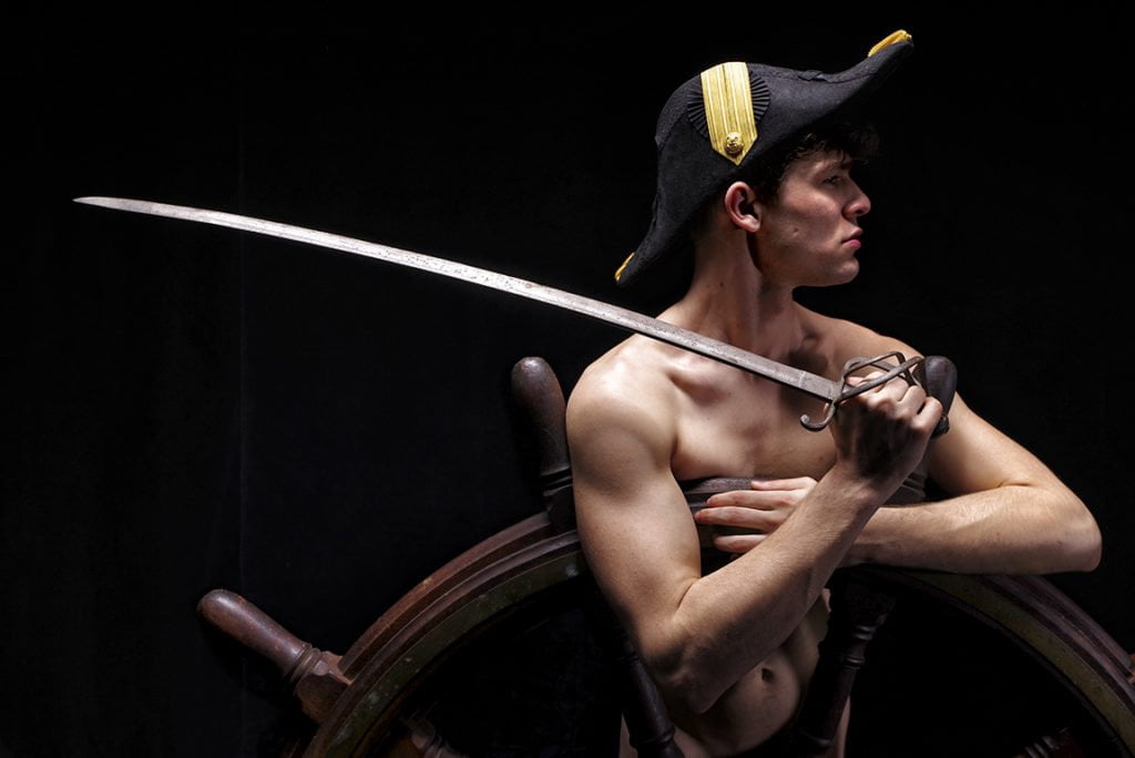 Nude man standing behind ships wheel with sword in hand