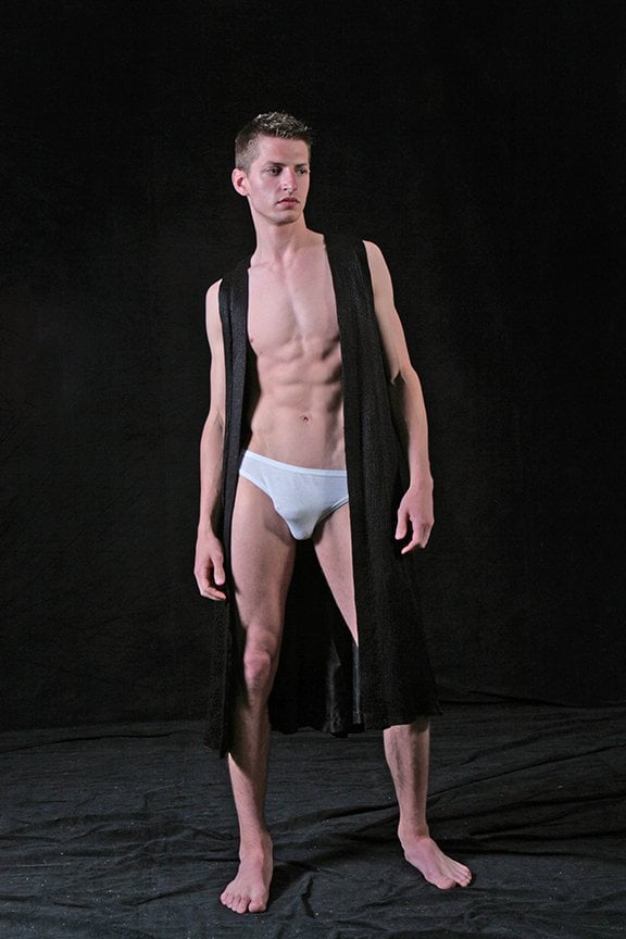 man wearing only underwear and a long vest in darkened studio