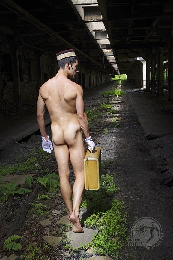 Naked man with bellhop hat and white gloves walking with suitcase in narrow beam of light in old train station