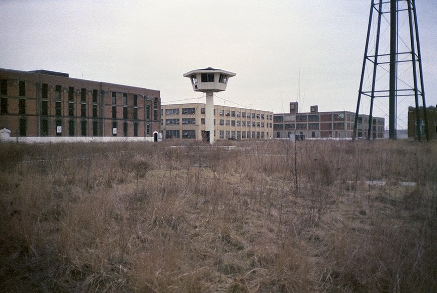 looking across field of weeds at buildings inside penitentiary wall, guard tower in center