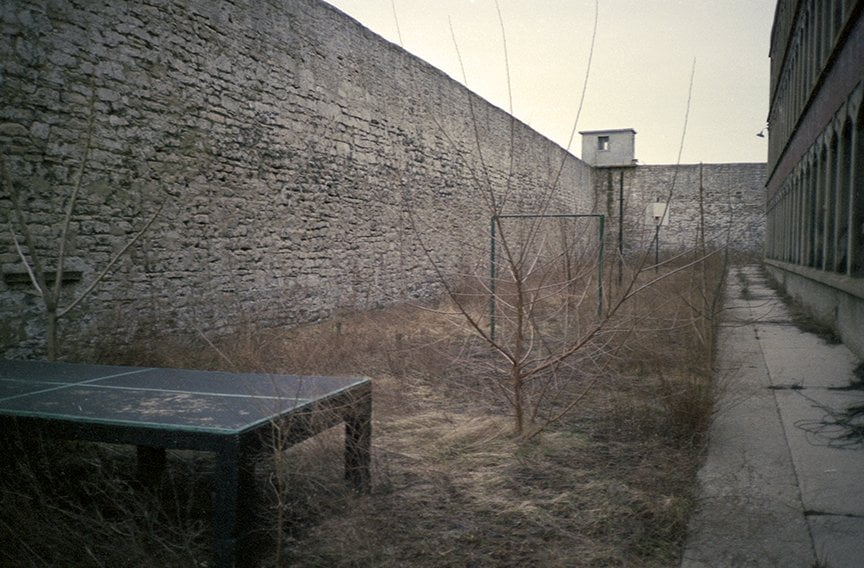 weed covered yard with large stone wall on left, small dormitory on right and abandoned pingpong table