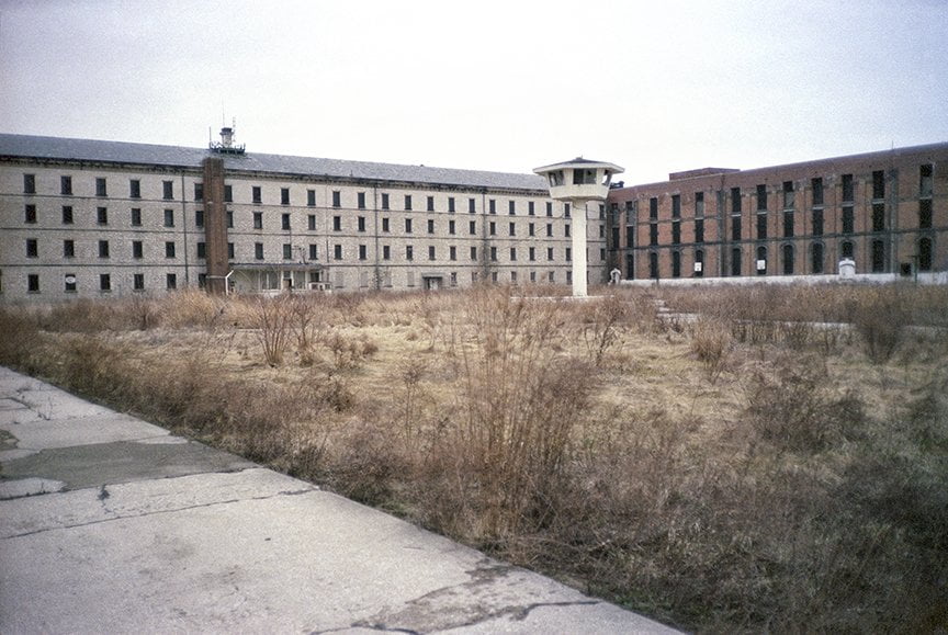 Another view of overgrown field with cellblock buildings and tower