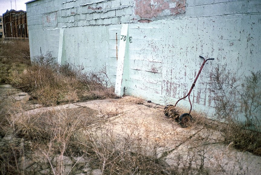 old push mower leaning against turquoise wall with weeds growing around it