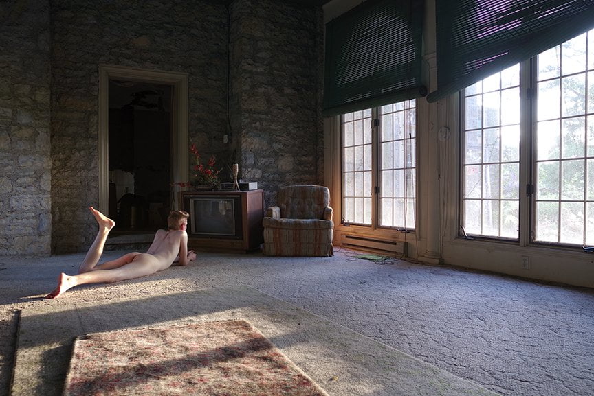 nude male laying on floor in front of old dead TV in room with big windows
