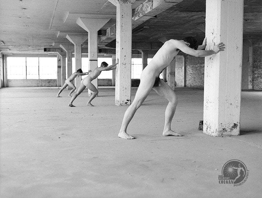 three nude men pushing on support pillars in empty warehouse