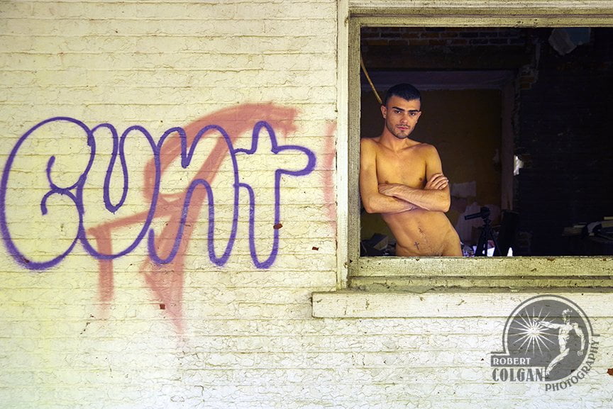 offset image of sirtless man standing in window of old house with profane graffiti spray painted on it