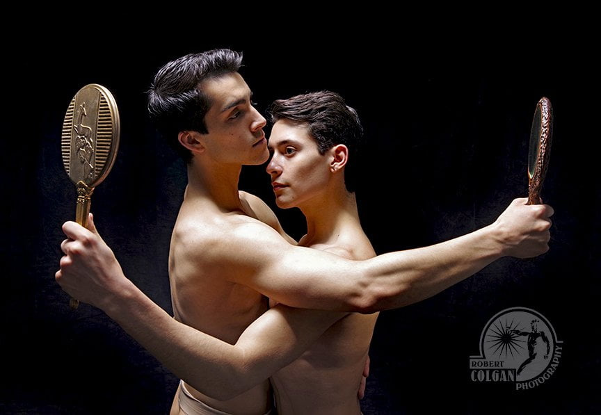 Two men embrace while looking past each other into hand held mirrors