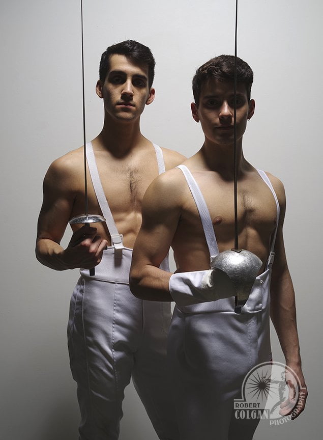 two men in white fencing pants look at the camera with swords in hand