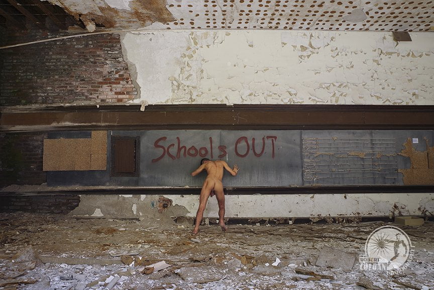 nude man pushing against remains of chalk board with "school's out" written on it