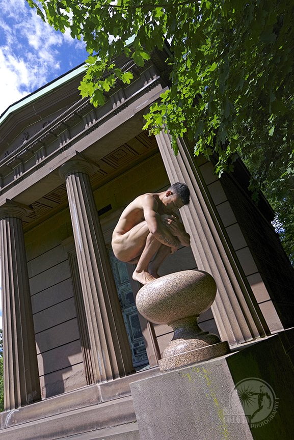 angular shot of nude man in fetal position standing on urn in front of classical structure