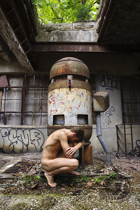 nude man hunkered down beside rusting machine