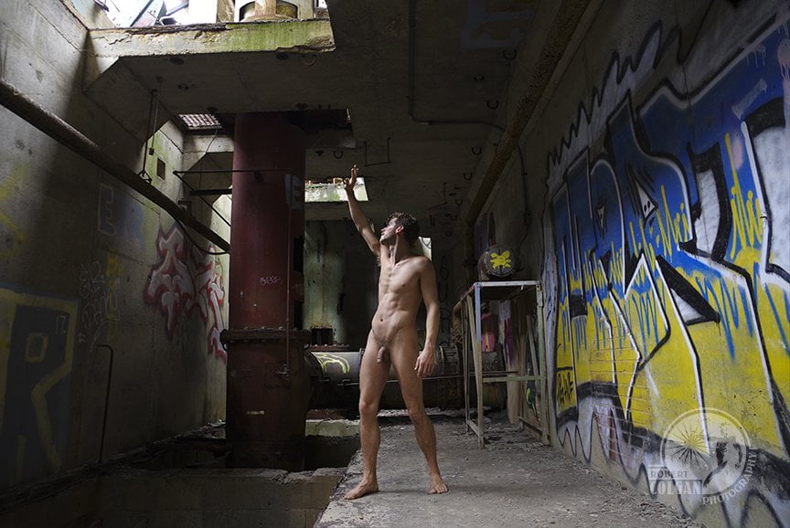 nude man standing in industrial room, hand raised to block light from above