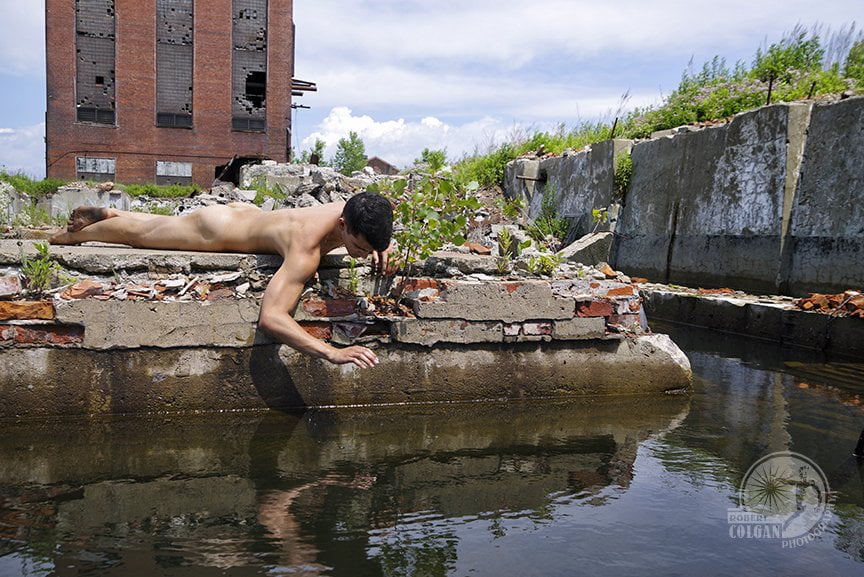 nude man lying on ground staring into dark waters in remains of old factory