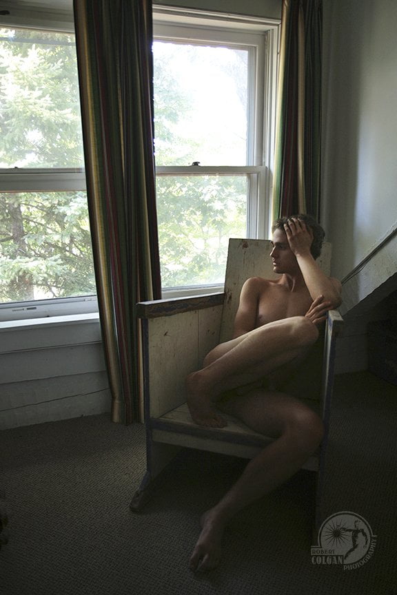 nude man curled up in wooden chair looking out window
