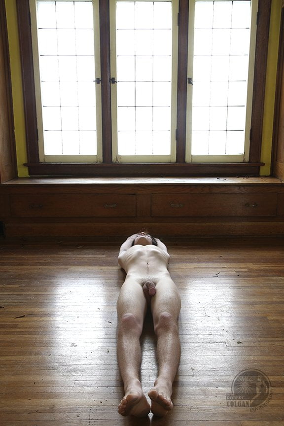 nude man lays pointing toward a bank of three windows