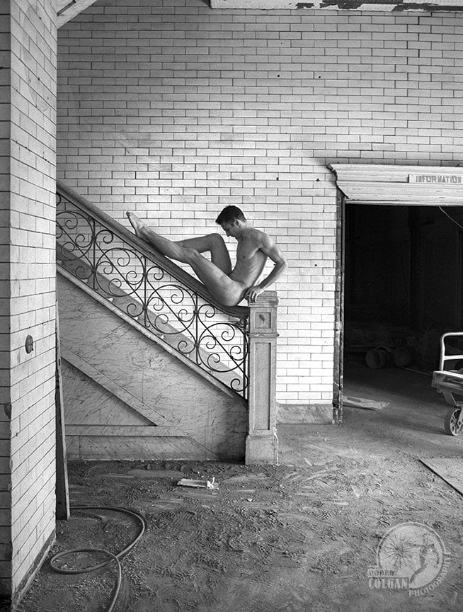 nude man sits on bannister of stairs feet pointing up