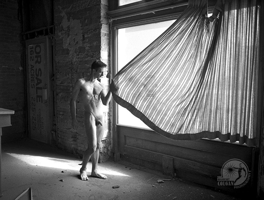 nude man in empty room pulling closed a curtain to outside window