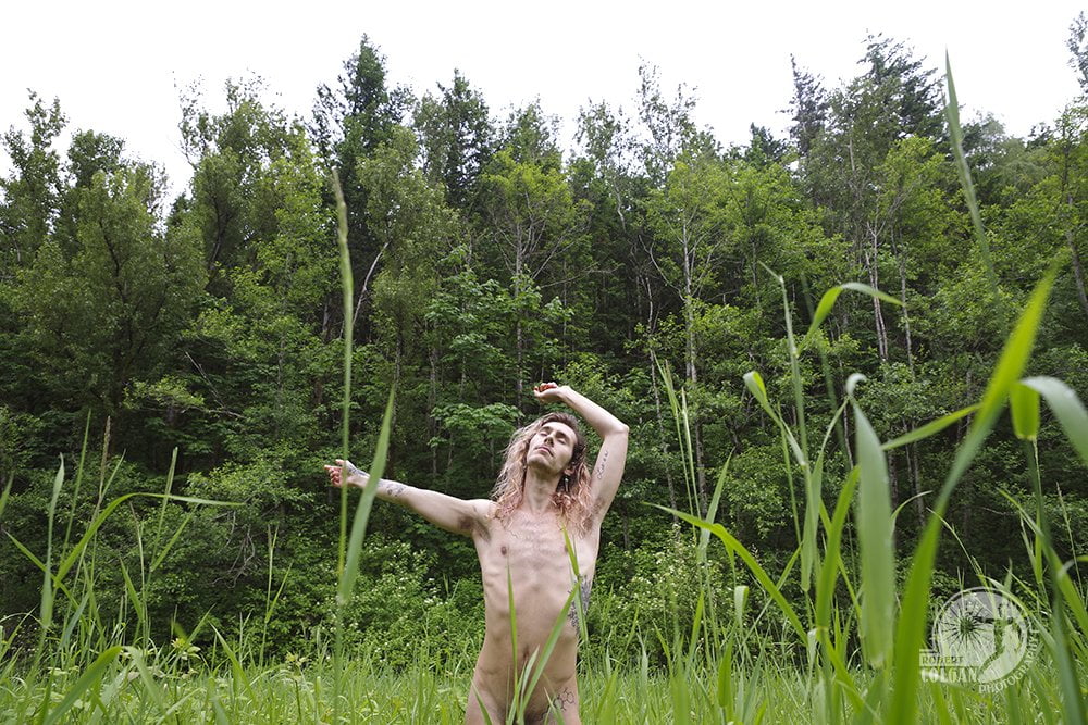 nude man with eyes closed in field with trees in background