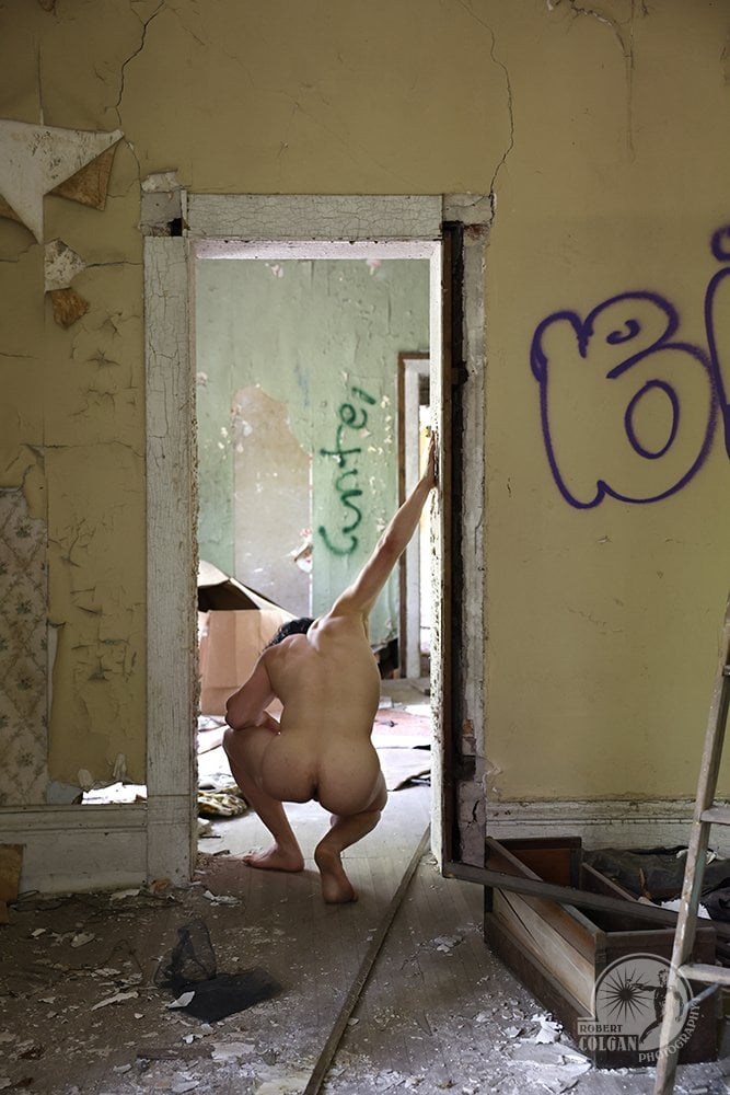 nude man with back to camera crouches in doorway of old house with hand extended up on frame