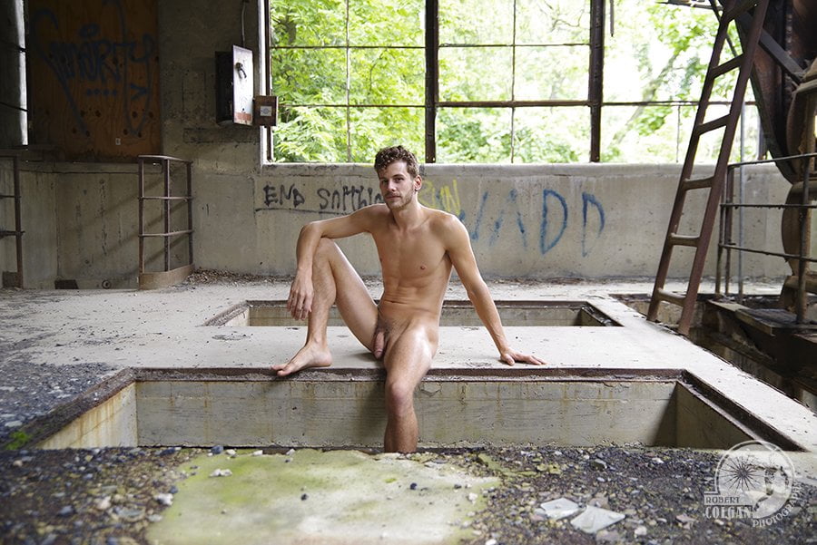 nude man sits in opening in concrete floor, leg dangling down