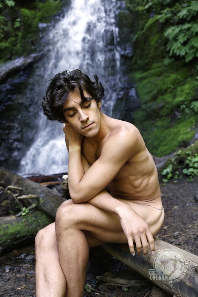 closeup of nude man sitting on log with waterfall in background