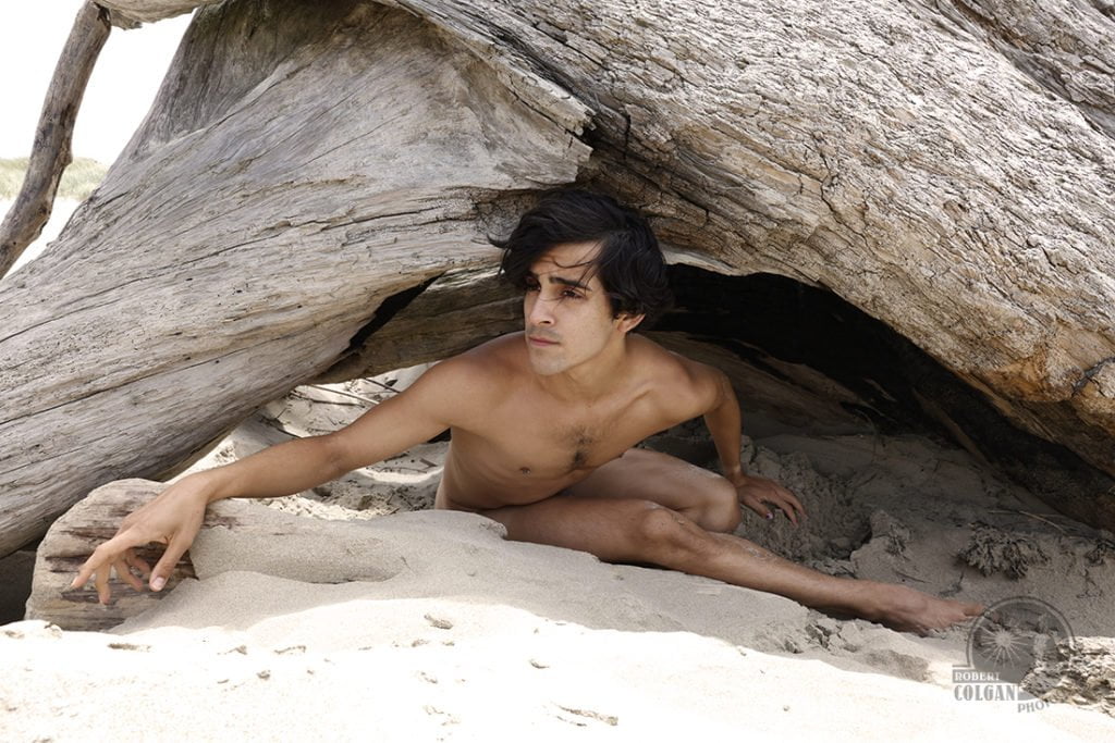 nude man looking out from beneath tree stump on beach