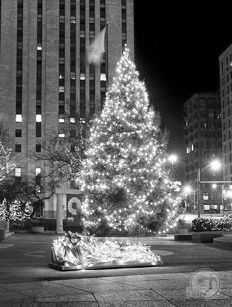 statue of Jesus wrapped in twinkle lights in front of outdoor Christmas tree