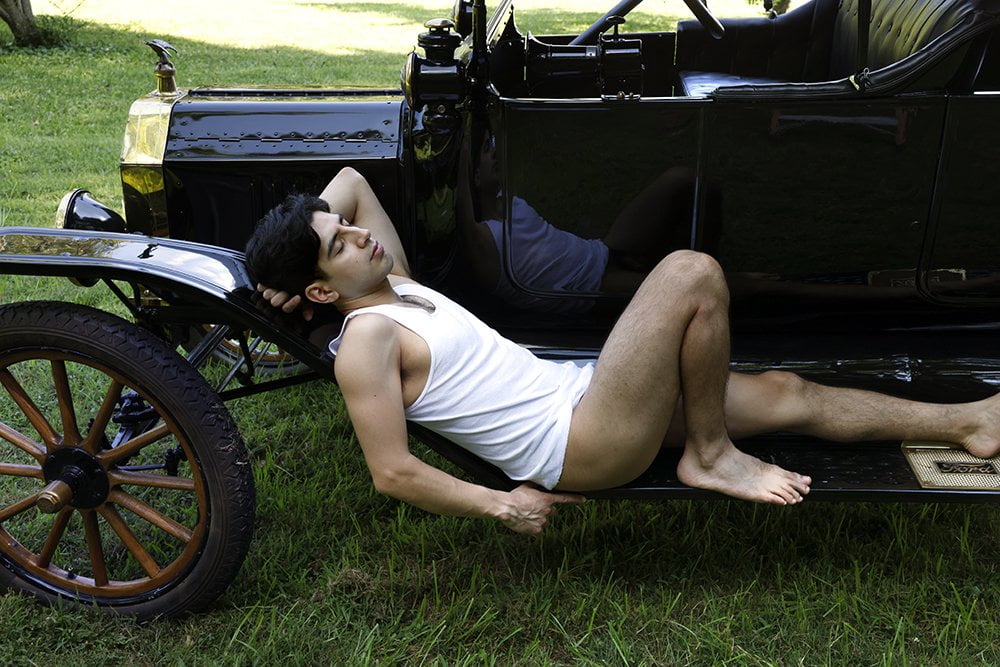 partially nude man reclines on fender of old car