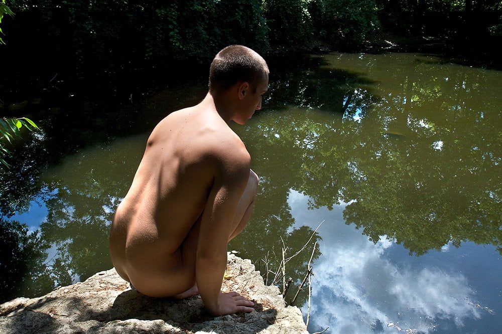 nude man sitting on rock overlooking pond