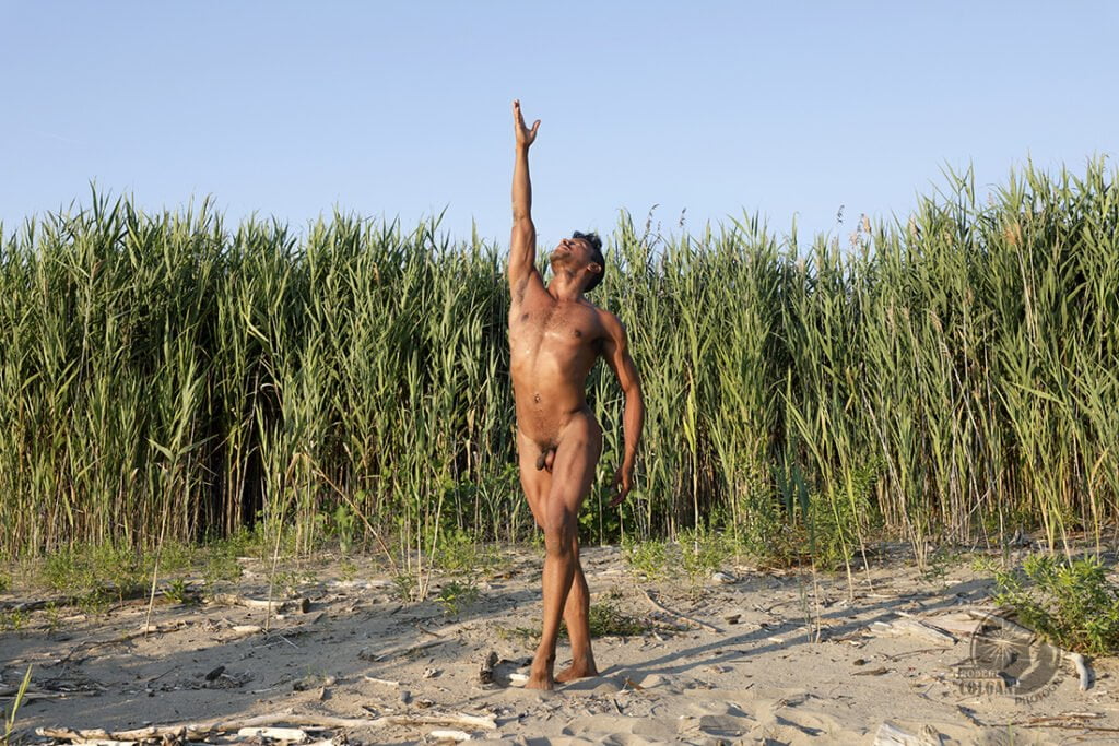 nude man reaches for sky with reeds in background