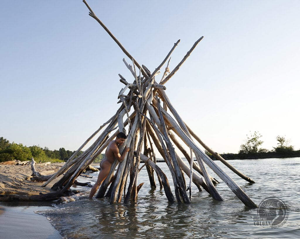 nude man standing beside tepee made of branches in lake