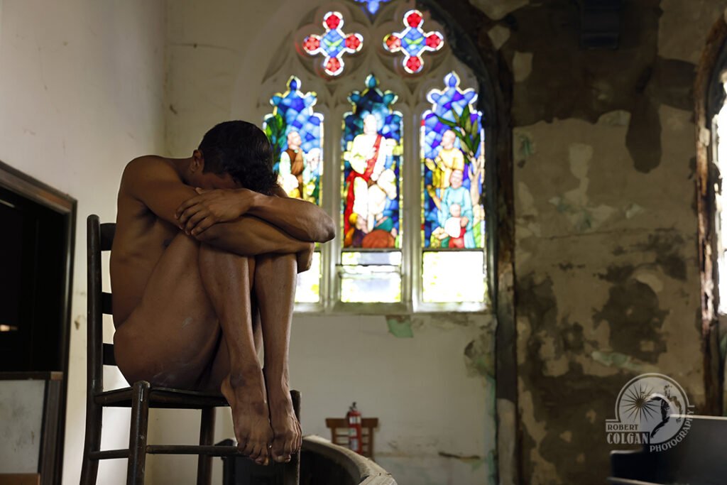 nude man curled up on chair with stained glass windows behind