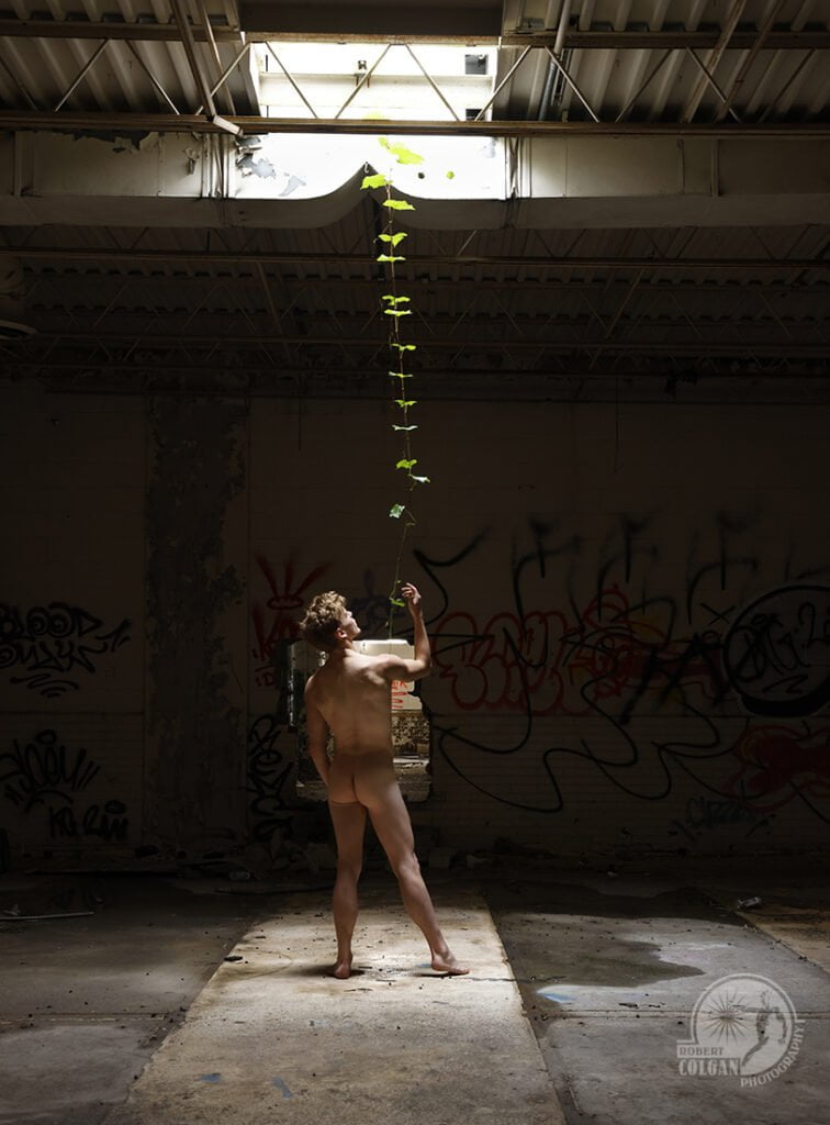 nude man in darkened room holding single vine dangling from ceiling