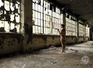 nude man stands in front of bank of windows of old factory
