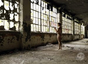 nude man stands in front of bank of windows of old factory