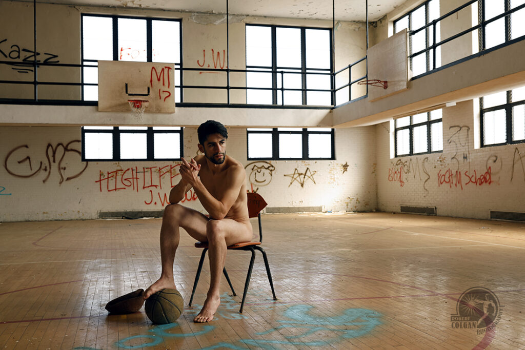 nude man shits on chair, foot on old basketball, in abandoned gymnasium