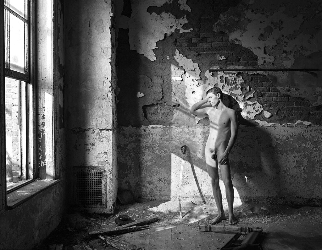 nude man standing in sunbeam next to dilapidated wall