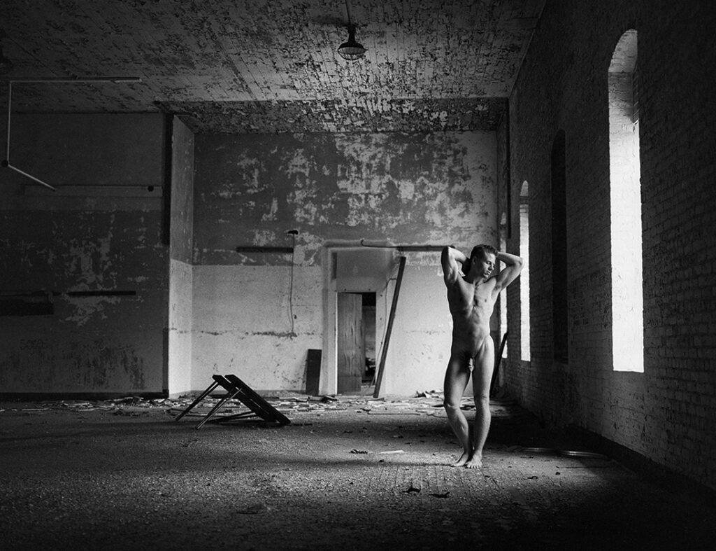 nude man stands with hands on head near window in deserted room