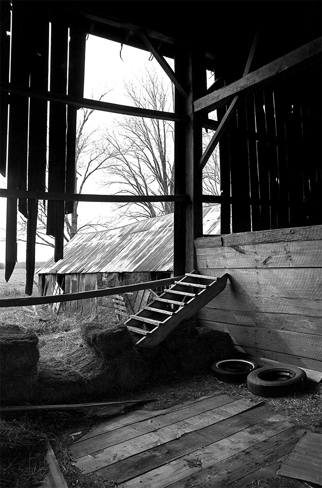 black and white image inside an old barn looking out at another old building