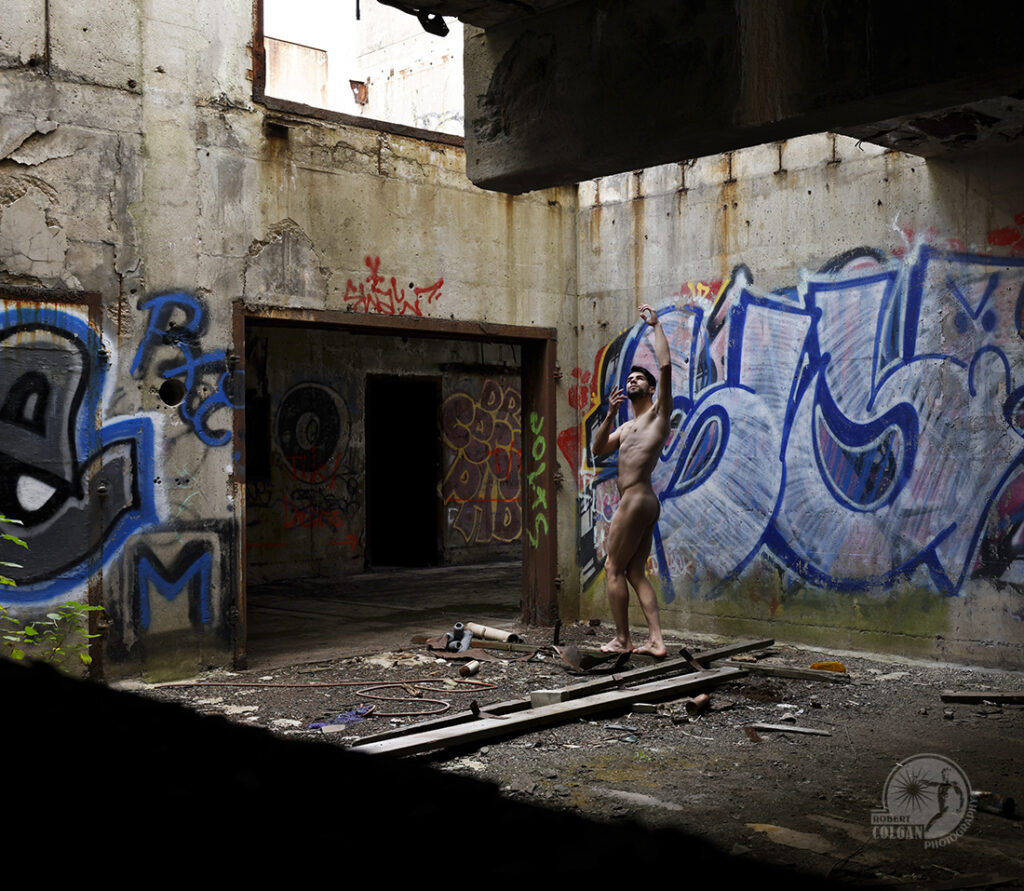 nude man raises arms toward skylight in abandoned factory