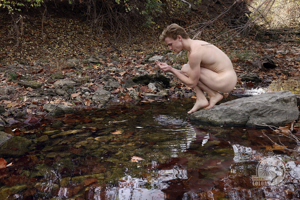 nude man perched on rock by stream