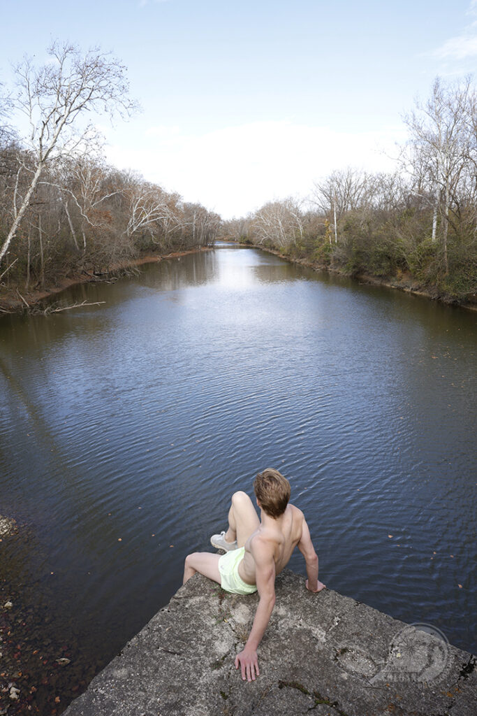 shirtless man sitting on bridge looking down river