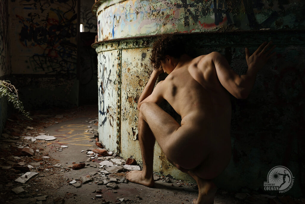 nude man crouching beside rusting metal container