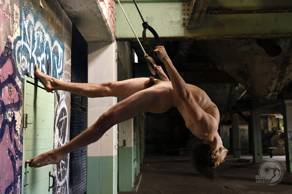 nude man shot from the side to demonstrate his acrobatic prowess on metal rings