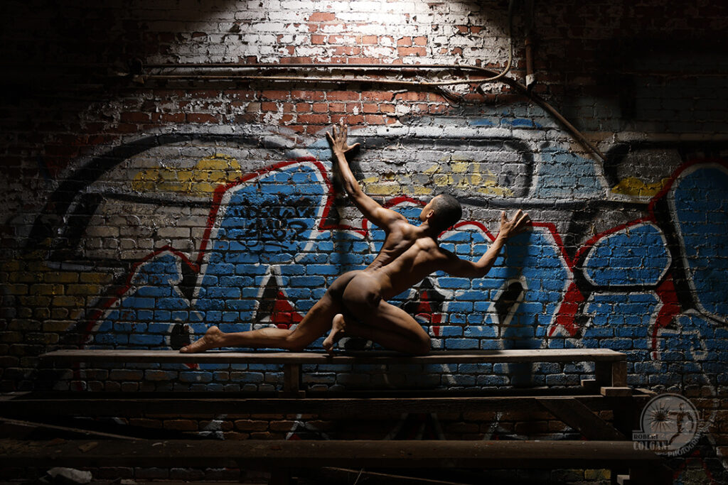nude man gestures against graffiti-covered wall with light streaming down from above