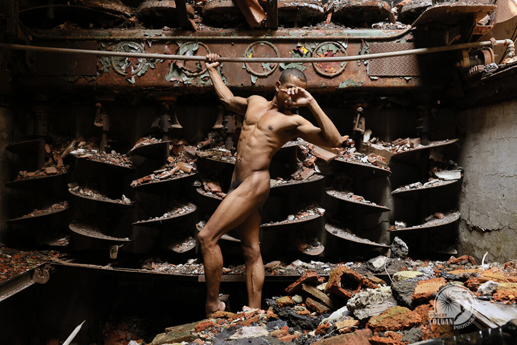 nude man stands beside a huge array of auger blades in old mill