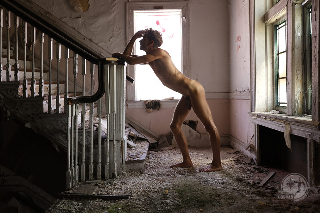 nude man leans against railing of rotting stairs in old church