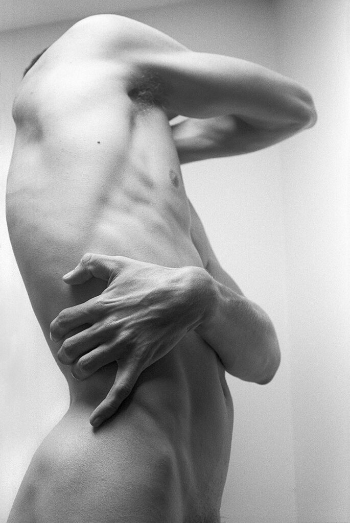 monochrome nude man photographed from the side as his arms grasp his body - one hand in close up