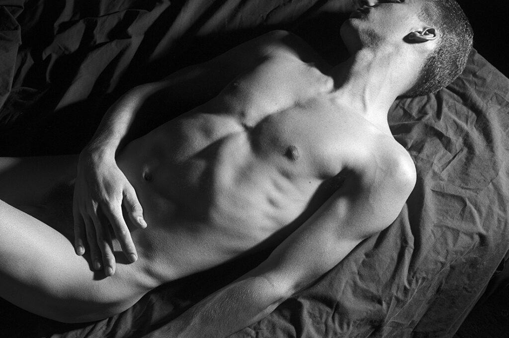 monochrome male nude photographed from above while laying on a bed