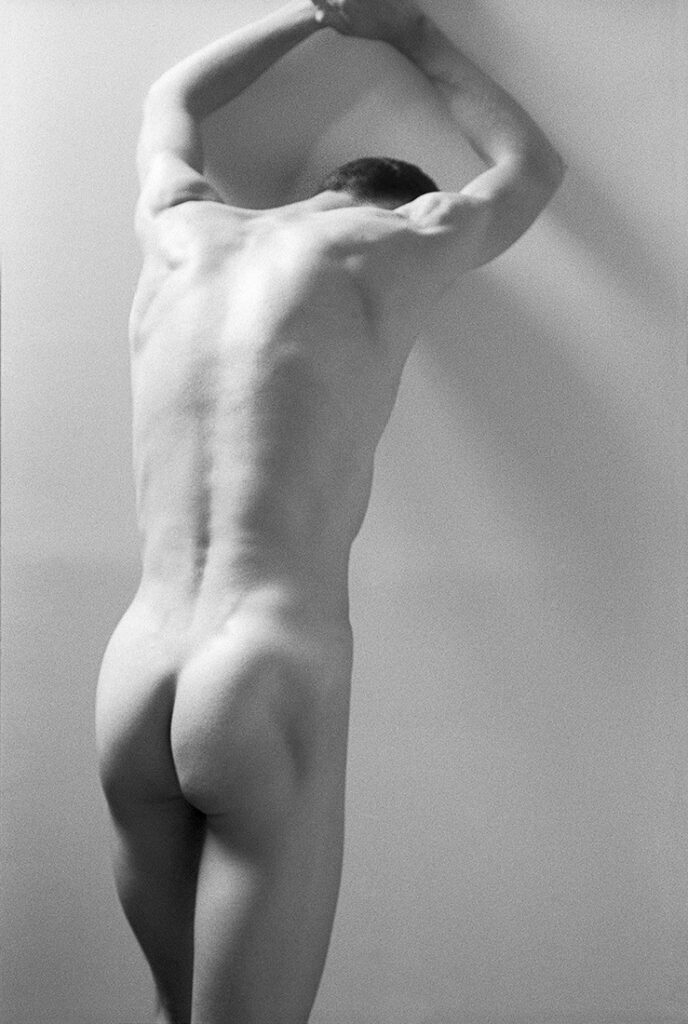 monochrome nude man photographed from behind as he braces himself against a wall